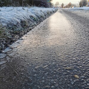 May include: A low-angle shot of a road surface covered in ice and water, reflecting the sky. The edge of the road is lined with frost-covered grass and snow. The scene suggests cold weather conditions.