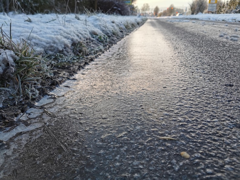 May include: A low-angle shot of a road surface covered in ice and water, reflecting the sky. The edge of the road is lined with frost-covered grass and snow. The scene suggests cold weather conditions.
