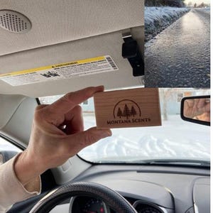 May include: A rectangular wooden air freshener with the text "MONTANA SCENTS" and a tree logo is held in a car. The car's interior is visible, including the dashboard, steering wheel, and sun visor. The background shows a snowy outdoor scene.