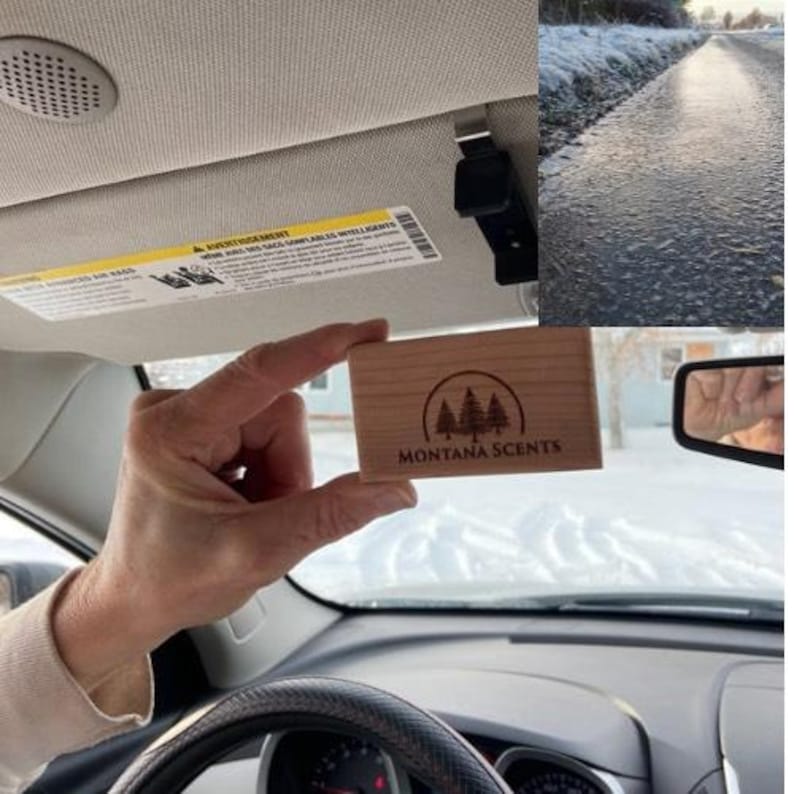 May include: A rectangular wooden air freshener with the text "MONTANA SCENTS" and a tree logo is held in a car. The car's interior is visible, including the dashboard, steering wheel, and sun visor. The background shows a snowy outdoor scene.