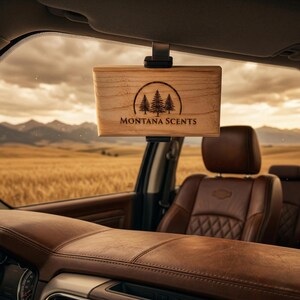 May include: A wooden car air freshener with the text "MONTANA SCENTS" and a tree graphic. The air freshener is hanging from the car's visor. The interior of the car is brown leather, with a view of a field and mountains in the background.