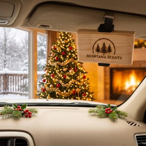 May include: A wooden car air freshener with the text "MONTANA SCENTS" and a tree logo is hanging from a car visor. The background shows a decorated Christmas tree and a fireplace, suggesting a holiday theme.