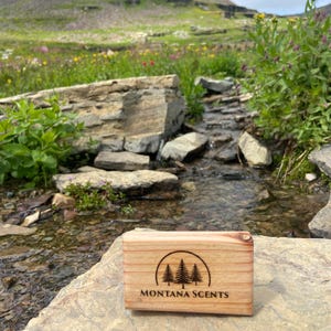 May include: A rectangular wooden box with the text "MONTANA SCENTS" and a tree logo is displayed on a rock. The box is light brown with visible wood grain. The background features a stream, rocks, and a mountain.