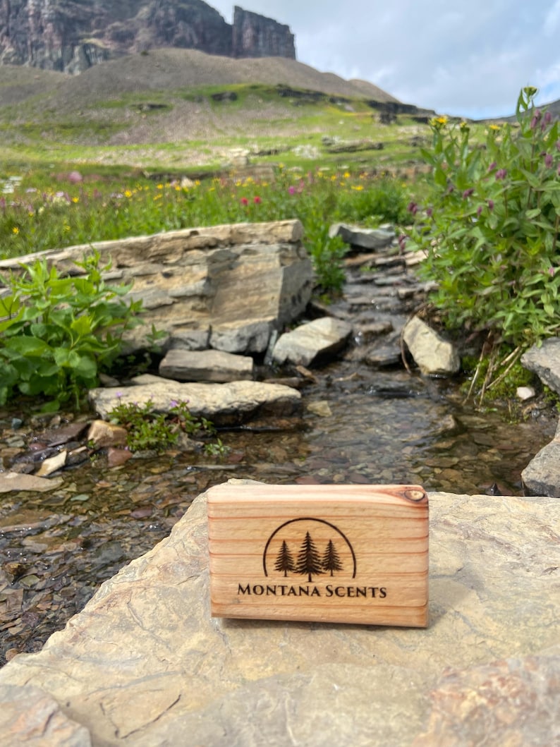 May include: A rectangular wooden box with the text "MONTANA SCENTS" and a tree logo is displayed on a rock. The box is light brown with visible wood grain. The background features a stream, rocks, and a mountain.