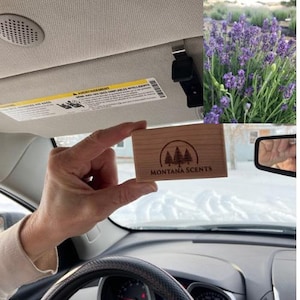 May include: A light-colored wooden air freshener with the text "MONTANA SCENTS" and a tree logo is held in a car. The car's interior is visible, including the dashboard, steering wheel, and a sun visor with a warning label. A sprig of lavender is in the background.