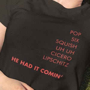 Pode incluir: T-shirt preto com texto vermelho. O texto inclui as frases "POP", "SIX", "SQUISH", "UH UH", "CICERO", "LIPSCHITZ" e "HE HAD IT COMIN'". A t-shirt é usada por uma pessoa deitada na relva verde.