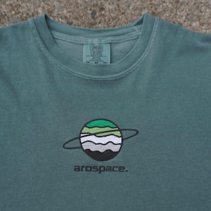 May include: A sage green t-shirt featuring an embroidered planet graphic with horizontal stripes in green, white, gray, and black, surrounded by a thin black ring. Below the planet, the word "arospace." is embroidered in black lowercase letters.