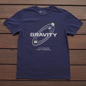 May include: A navy blue short-sleeve t-shirt featuring a white graphic of three orbiting planets and the text GRAVITY. Below the graphic, the text reads: is my favorite force of attraction. The shirt has a crew neckline and a standard fit.