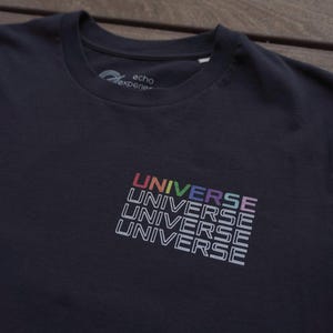 May include: A dark charcoal gray crew neck t-shirt featuring a graphic print on the chest. The word UNIVERSE is stacked four times in white outline lettering, with the top row featuring a multicolored gradient effect across the letters.