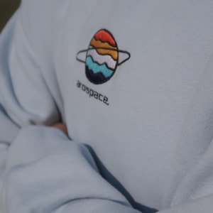 May include: Pale blue sweatshirt featuring an embroidered planet design in red, orange, yellow, white, blue, and black. The word "arospace" is embroidered below the planet. The sweatshirt is made of a soft material.