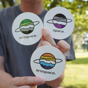 Könnte beinhalten: Drei runde weiße Aufkleber mit Planetenillustrationen und Ringen. Ein Aufkleber zeigt den Text arospace mit einem grünen, weißen und grauen Planeten. Ein weiterer zeigt space mit einem grauen, weißen und violetten Planeten. Der dritte zeigt arospace mit einem orangefarbenen, gelben, weißen und blauen Planeten.