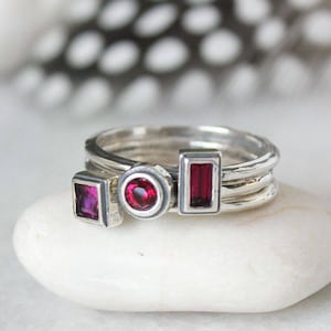 May include: A set of three silver rings with red gemstones. The rings are stacked on top of each other and feature different shaped gemstones.