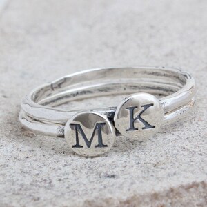 May include: Two silver rings with a round disc on each ring. The disc on the left ring has the letter "M" engraved on it. The disc on the right ring has the letter "K" engraved on it.