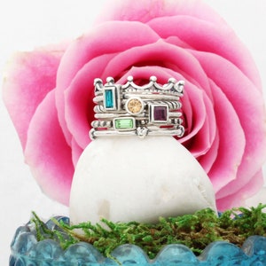 May include: A set of silver rings with colorful gemstones, stacked on a white stone and surrounded by green moss. The rings feature a crown design and various colored stones, including turquoise, green, and purple.