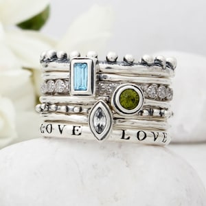 May include: A stack of sterling silver rings with various gemstones, including a rectangular light blue stone, a round green stone, and a marquise-cut clear stone. Some rings have small clear stones and the word "LOVE" engraved on them.