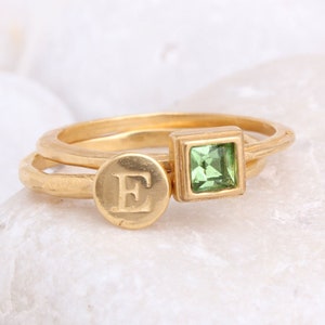 Puede incluir: Dos anillos de oro. Uno presenta una gema verde claro cuadrada. El otro tiene un diseño circular con la letra "E" grabada. Los anillos están sobre un fondo blanco.