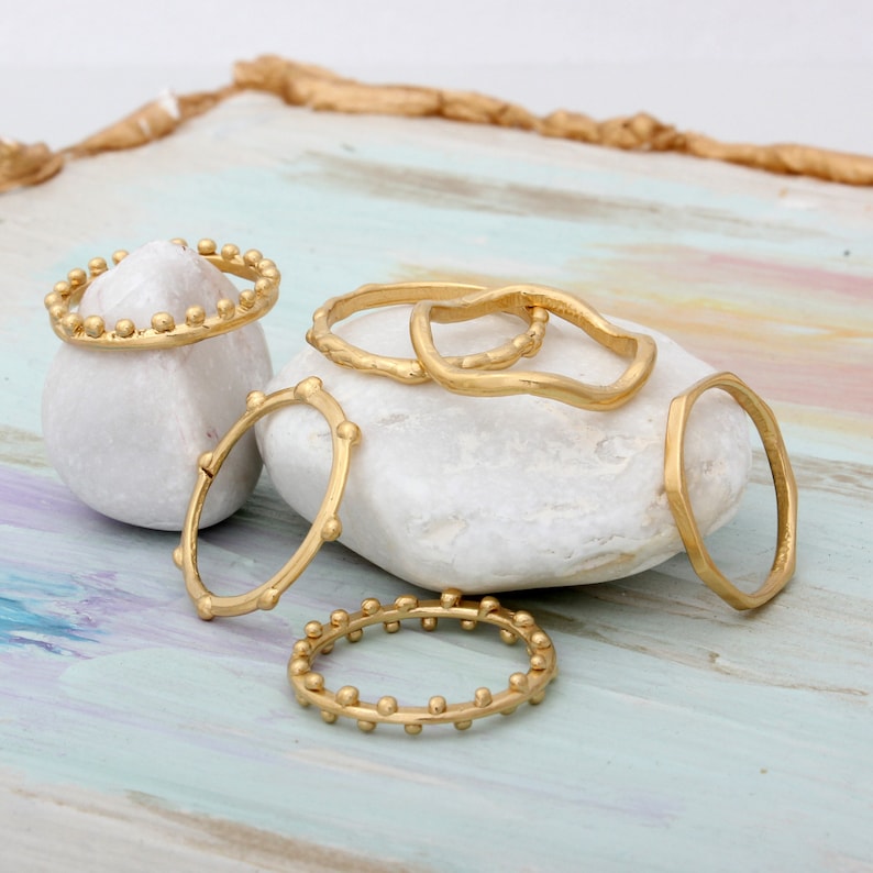 May include: Set of five gold-toned rings with different designs. The rings are stacked on a white stone and have a textured, hammered finish.