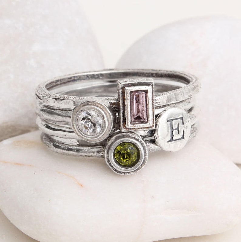May include: A set of five silver rings with different stones and an initial "E". The rings are stacked on top of each other and have a hammered texture.