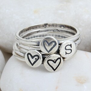 May include: Four silver stacking rings with a heart design and the letter 'S' engraved on the top. The rings are stacked on top of each other.