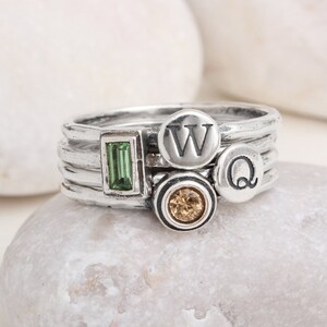 May include: A set of four silver stacking rings with a green rectangular stone, a brown round stone, and two letter charms, one with the letter "W" and the other with the letter "Q".