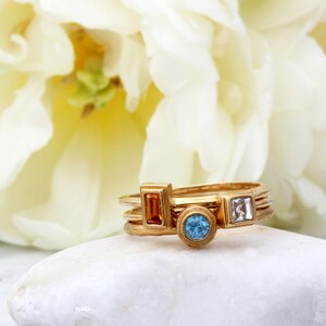 May include: Gold ring set with three gemstones. The ring features a rectangular orange stone, a round blue stone, and a square clear stone.
