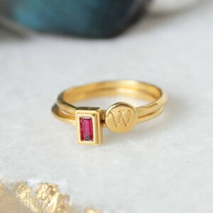 May include: A gold ring with a small red gemstone and a round charm with the letter "W" engraved on it.