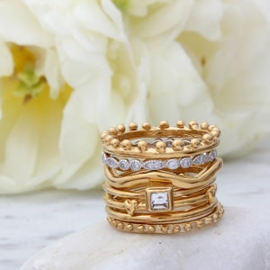 May include: A stack of gold rings with a variety of textures and designs, including a ring with a square-shaped clear stone.