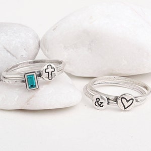 May include: Three silver rings with different charms. The first ring has a turquoise stone, a cross, and a silver band. The second ring has a heart charm and a silver band. The third ring has an ampersand charm and a silver band.