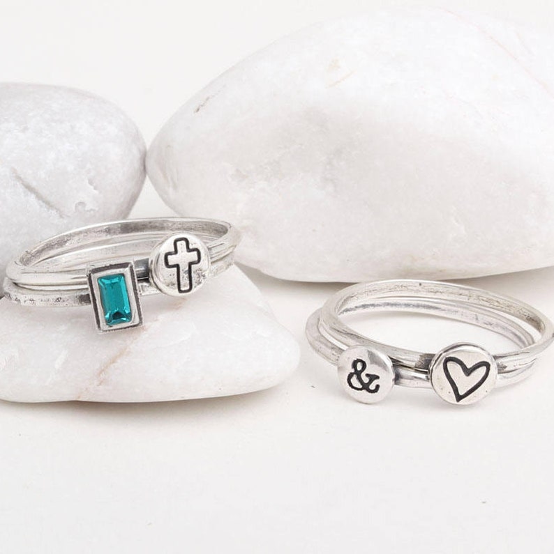 May include: Three silver rings with different charms. The first ring has a turquoise stone, a cross, and a silver band. The second ring has a heart charm and a silver band. The third ring has an ampersand charm and a silver band.