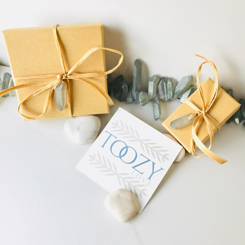 May include: Two small, brown gift boxes tied with twine and decorated with a crystal point. The boxes are surrounded by clear quartz crystals and a white card with the text "Toozy Love" in blue.