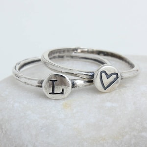 May include: Two silver rings, one with a heart and the other with the letter L, stacked on top of each other.