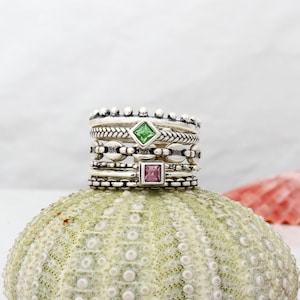 May include: A stack of five silver rings with a green and a pink square gemstone. The rings are on a textured, light green surface.