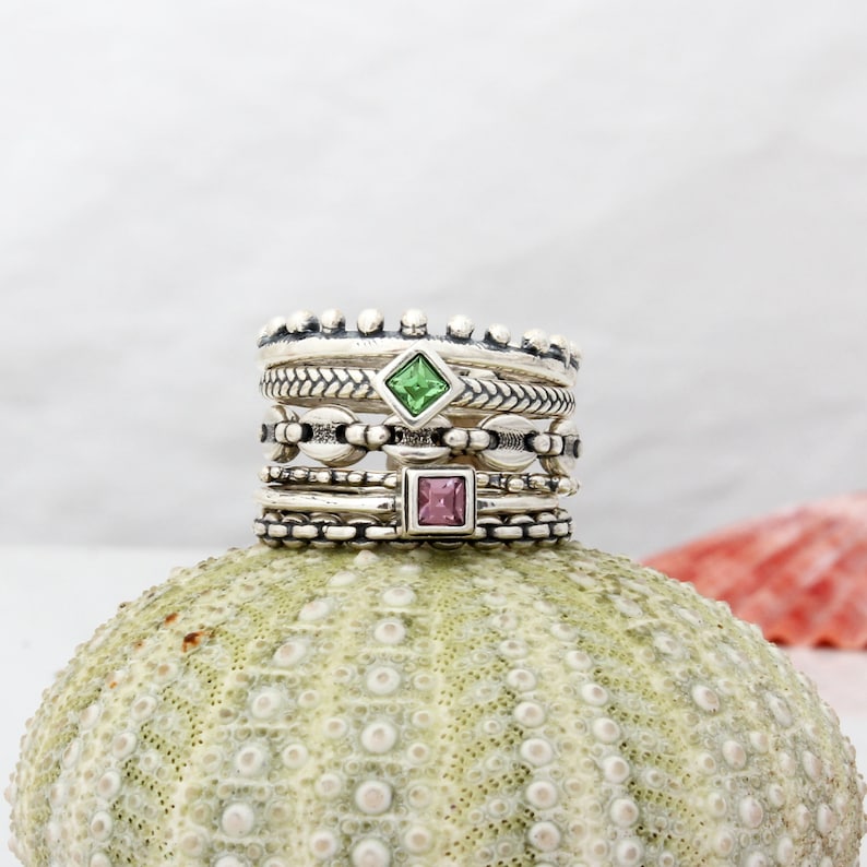 May include: A stack of five silver rings with a green and a pink square gemstone. The rings are on a textured, light green surface.
