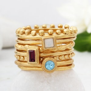 May include: A set of seven gold rings with different colored gemstones. The rings are stacked on top of each other and feature a variety of textures, including smooth, textured, and beaded.
