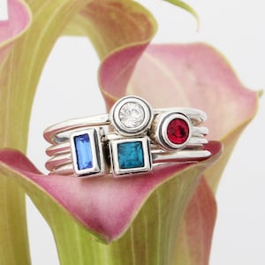 May include: A set of four silver rings with colorful gemstones. The rings feature a round clear stone, a square blue stone, a rectangular blue stone, and a round red stone.