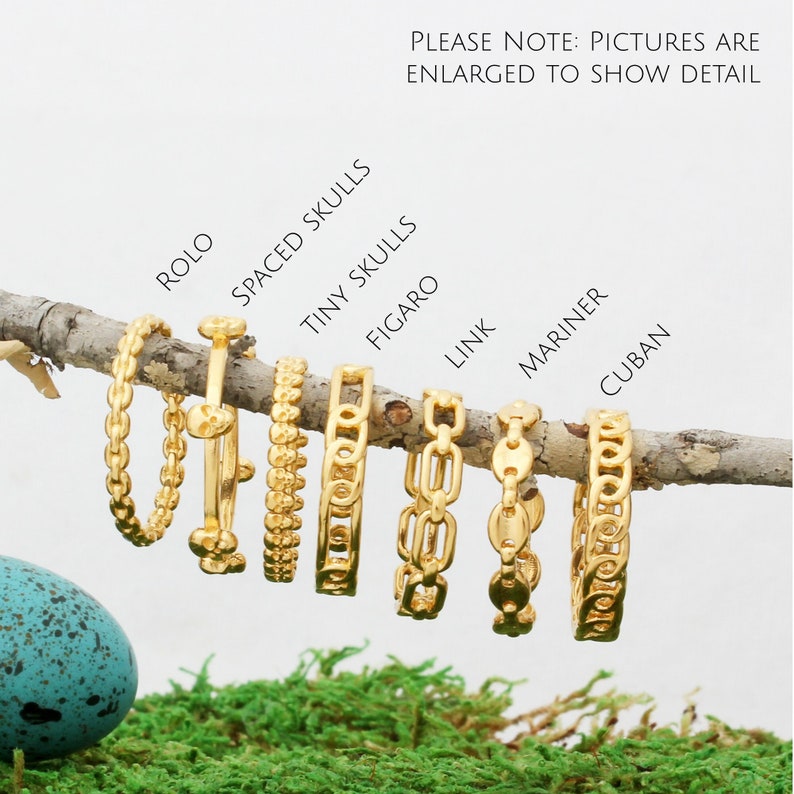 May include: A close-up of eight gold chains of different styles, including rolo, spaced skulls, tiny skulls, figaro, link, mariner, and cuban. The chains are arranged in a row on a branch with a green moss background.