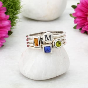 May include: Four silver stacking rings with colorful gemstones. The top ring has a round silver charm with the letter "M" on it. The other rings have square gemstones in orange, blue, and green.