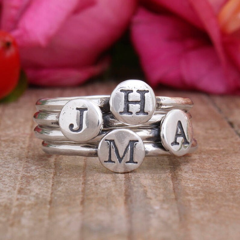 May include: Four silver rings with initial letter charms. The rings are stacked on top of each other and the charms are visible. The charms spell out the letters "J", "M", "A", and "H".