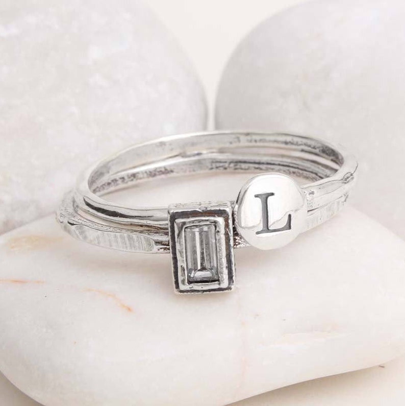 Pu&ograve; includere: Un anello d'argento con tre bande, una con una pietra trasparente rettangolare, una con una superficie testurizzata e l'ultima con la lettera "L" incisa su un disco rotondo.