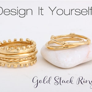 May include: A set of gold stack rings, featuring a variety of textures and designs. The rings are stacked on a white stone. The text "Design It Yourself!" and "Gold Stack Rings" are visible in the image.