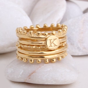 May include: Gold-toned stackable rings with a textured finish. The top ring features a round disc with the letter "K" engraved on it.