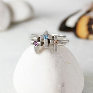 May include: Three silver rings with small cross charms. Each cross has a different colored gemstone in the center: blue, pink, and orange.