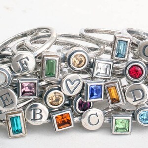 May include: A collection of silver rings with various colored gemstones and letter charms. The rings feature a variety of shapes, including circles, squares, and rectangles. The gemstones are set in silver bezels and are surrounded by a black border.