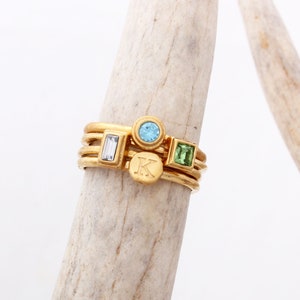 May include: Gold stacking rings with a blue, green, and initial "K" charm. The rings are on a wooden surface.
