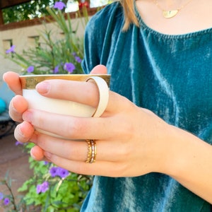 May include: A person wearing a teal blue velvet shirt holds a white teacup with a gold rim. The person is wearing a gold ring with small white stones on their ring finger.