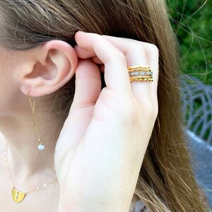 May include: A close-up of a person's hand wearing three gold rings. The rings are stacked on top of each other and feature a variety of designs, including a beaded ring, a ring with small diamonds, and a simple band.