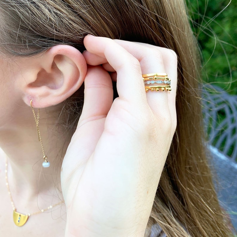 May include: A close-up of a person's hand wearing three gold rings. The rings are stacked on top of each other and feature a variety of designs, including a beaded ring, a ring with small diamonds, and a simple band.