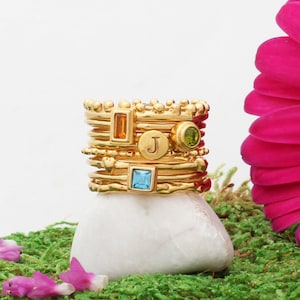 Puede incluir: Una pila de anillos de oro con diferentes piedras preciosas de colores, incluyendo una piedra cuadrada azul, una piedra redonda verde y una piedra rectangular naranja. El anillo superior tiene la letra "J" grabada.