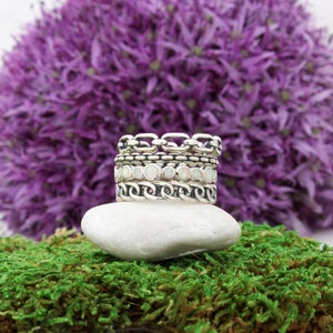 May include: A set of four silver rings stacked on a white rock. The top ring is a chain link design, the second ring is a band with small white opal stones, the third ring is a chain link design, and the bottom ring is a plain band. The rings are set against a background of purple flowers and green moss.