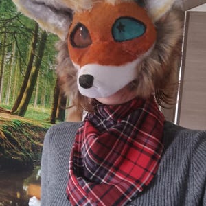 Può includere: Una testa di fursuit di volpe con pelliccia arancione, marrone e bianca, con grandi occhi espressivi. È abbinata a una sciarpa a quadri rossa e nera e a un maglione grigio.